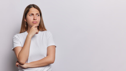 Horizontal shot of pensive long haired woman keeps hand on chin concentrated aside being thoughtful dressed in casual t shirt isolated over white background with copy space for your promotion