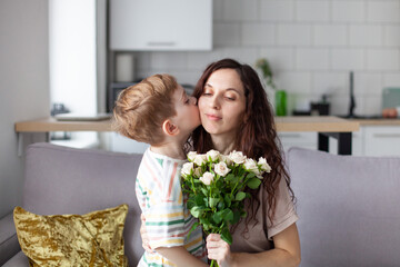 Little boy kissing his mother and giving her a bouquet of roses at home. Mother's day concept. Selective focus.