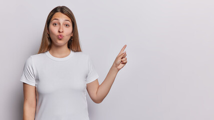 Horizontal shot of pretty long haired woman keeps lips rounded and points index finger on blank space attracts your attention to something wears casual t shirt isolated over white background