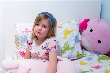 Small girl sitting on bed set with dinosaur patterned sheets surrounded by large stuffed toy cushions looking forlorn