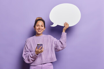 Horizontal shot of pleasant looking European woman uses mobile phone for chatting online holds communication bubble for your advertising content smiles broadly isolated over purple background