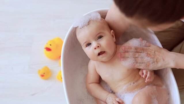 A cute little big-eyed beautiful baby is bathing in warm water with bubble bath in the hands of a caring mother in a small tub. The concept of child care.