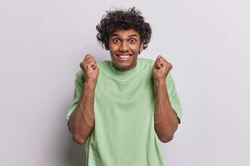 Joyful curly haired Hindu man feels ecstatic keeps fists raised up celebrates good luck smiles broadly dressed in casual green t shirt poses against white background. People joy and happiness concept