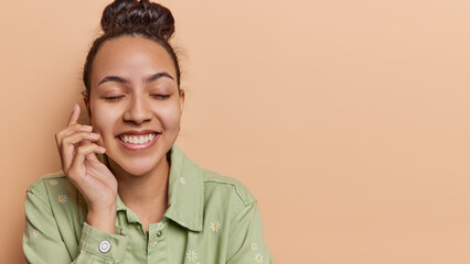 Horizontal shot of pretty pleased woman keeps eyes closed smiles toothily recalls something pleasant wears green jacket isolated over brown background copy space for your promotional content