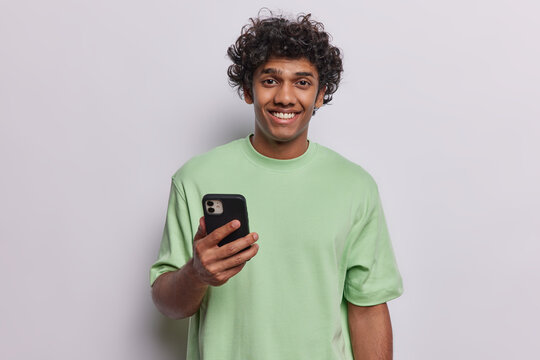 Studio shot of handsome curly Hindu man uses mobile phone for communication and staying in touch smiles broadly dressed in casual green t shirt isolated over white background. Technology concept