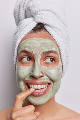 Vertical shot of cheerful dreamy woman applies beauty clay mask bites finger concentrated aside poses with bath towel on head isolated over white background. People wellness and facial treatments