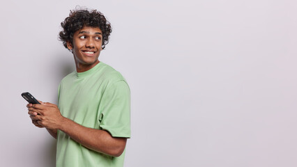 Horizontal studio shot of handsome Hindu millennial guy browsing social networks and messaging checking new app on modern cellphone focused back dressed in green t shirt isolated over white background