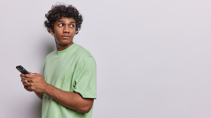Horizontal shot of serious curly haired Hindu man uses mobile cell phone chatting and sending sms looks back notices someone dressed in green t shirt isolated over white background copy space for text