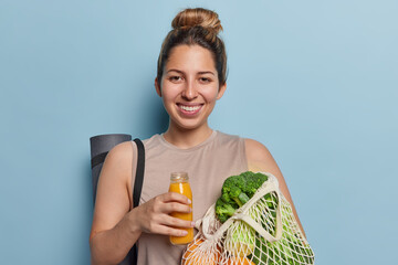 Horizontal shot of cheerful European woman with hair bun smiles broadly holds glass bottle of fresh orange juice and net bag full of vegetables dressed in t shirt isolated over blue background
