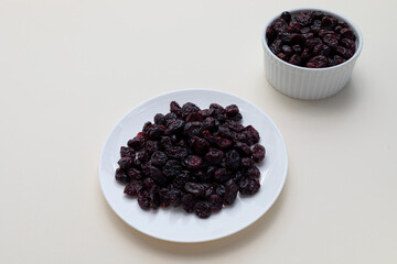 Fototapeta na wymiar Dried Cranberries On White Plate On Beige Background. Superfood. Selective Focus. Sweet Dehydrated Cranberries. Nutrients And Vitamins. Horizontal Plane.