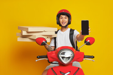 Happy oriental boy in red helmet sits on scooter on yellow background, holds pizza boxes in one hand and phone in another, fast delivery concept, copy space