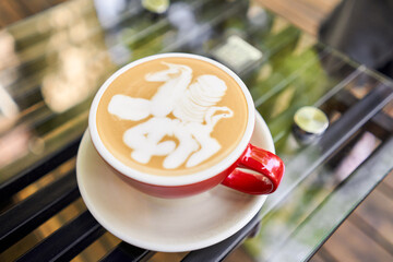 Cup of hot cappuccino with Pegasus figurine on milk foam. The table in the coworking space of the coffee shop. A cup of coffee on the veranda on a sunny day. Coffee break at the cafe. Close-up