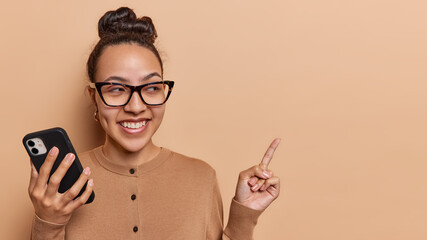 Happy dark haired woman wears optical eyeglasses and casual jumper points index finger on copy space for your advertising content suggests to check this out uses mobile phone for chattning online