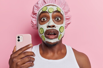 Photo of shocked dark skinned man takes selfie via smartphone holds breath from amazement applies facial clay mask with soothing cucumber slices wears bath hat and white t shirt isolated on pink wall