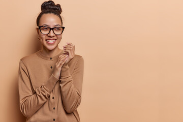Horizontal shot of pleased pretty woman with hair bun smiles gladfully focused aside rubs hands wears optical eyeglasses and casual jumper isolated over brown background copy space for your text