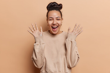 Horizontal shot of cheerful Latin woman with hair bun exclaims loudly keeps palms raised up being in good mood dressed in casual jumper isolated over brown background. People and happiness concept