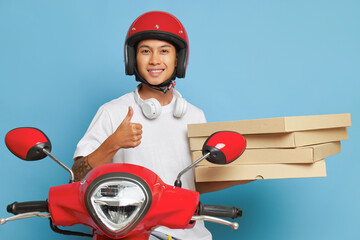 Happy asian boy in red helmet sits on scooter on blue background in studio, holds pizza boxes and smiles widely, fast delivery concept, copy space © South House Studio