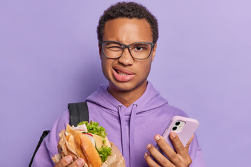 Discontent dark skinned man winks eye looks unhappily at camera holds sandwich and smartphone feels dissatisfied dressed in casual hoodie isolated on purple background. Fast food and nutrition concept