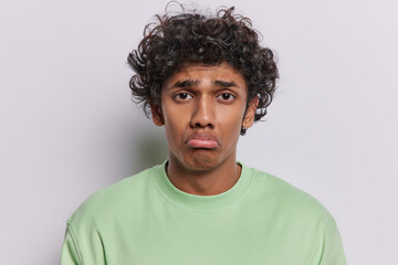 Portrait of frustrated disappointed curly haired Hindu man purses lips has upset offended expression looks at camera sadly frowns eyebrows dressed in green t shirt isolated over white background.