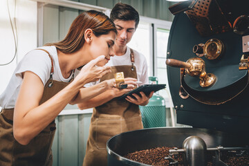 Young Adult Small Business Owner's Caucasian Couple Woking Together in Coffee Roaster Factory, Professional Barista Checking the Quality and Aroma After Roasted with Machine in Warehouse.