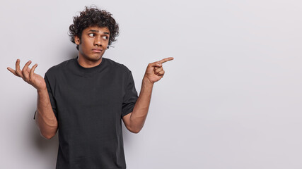 Confused handsome Hindu man with curly hair shrugs shoulders with hesitation and points index finger at empty space feels doubtful dressed in casual black t shirt isolated over white background