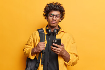 Horizontal shot of serious curly haired Hindu man concentrated in smartphone reads news in internet wears casual shirt poses with headphones around neck carries rucksack isolated on yellow background