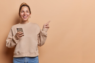 Horizontal shot of pretty cheerful woman smiles broadly uses mobile phone for online communication dressed in casual pullover and jeans isolated over brown background. Technology and advertisement