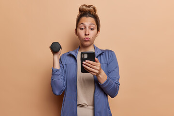 Sporty young woman with hair bun raises dumbbell focused in smartphone has stunned expression reads news while doing sport wears sports jacket isolated over brown background. Healthy lifestyle