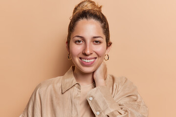 Portrait of cheerful young woman keeps hand on neck smiles broadly wears shirt and golden earrings looks directly at camera isolated over brown background. People and happy emotions concept.