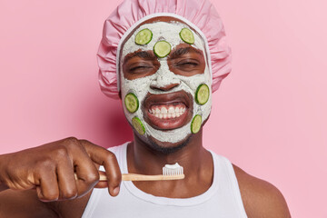 Horizontal shot of dark skinned man with cucumber slices on face holds toothbrush with toothpaste cleans teeth keeps eyes closed undergoes daily hygiene treatments isolated over pink background