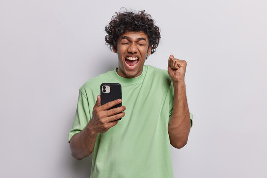 Overjoyed Curly Hindu Middle Aged Man Using Smartphone Making Winner Gesture And Exclaims Loudly Celebrating Win Shaking Clenched Fist Dressed In Green T Shirt Isolated Over White Background