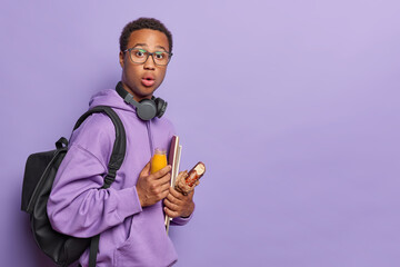 Sideways shot of scared dark skinned man holds bottle of fresh juice and bun textbooks wears spectacles hoodie carries rucksack isolated over purple background copy space for your promotional content