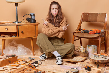 Hesitant clueless woman carpenter points sideways tries to choose something dressed in workwear feels confused poses on floor in carpentry shop with sawdust surrounded by different instruments