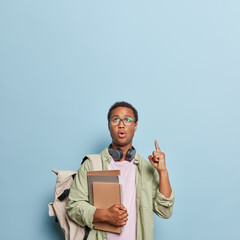 Vertical shot of dark skinned adult man points index finger overhead stands speechless carries textbooks and backpack demonstrates place for your advertisement isolated over blue background.