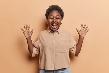 Young cheerful african girl wearing brown blouse and blue jeans with short curly hair and two raised hands happy to receive good news. Isolated on brown background expressing positive emotions