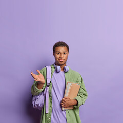 Vertical shot of hesitant dark skinned millennial guy shrugs shoulders feels confused carries backpack on shoulder carries notepads being clueless isolated over purple background copy space overhead
