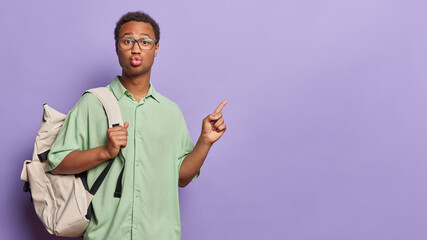 Horizontal shot of handsome dark skinned man keeps lips folded points at blank copy space for your advertising content stands with rucksack isolated over purple background shows educational content
