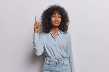 Horizontal shot of curly haired woman points index finger above demonstrates something on ceiling gives recommendations wears blue shirt and jeans isolated over white background. Follow this direction