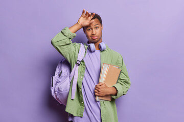 People and education concept. Tired dark skinned skilled male student keeps hand on forehead feels exhausted carries rucksack and textbooks necessary for studying isolated over purple background