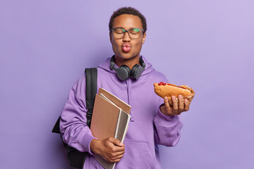 Photo of dark skinned teenage guy keeps lips folded eats tasty hotdog after having classes carries notepads dressed in sweatshirt isolated over purple background. Fast food is hamrful for your health