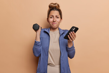 Indoor shot of hard working sportswoman exercises with dumbbell downloads app for sport on smartphone dressed in activewear isolated over brown background. Pilates fitness healthy lifestyle concept