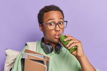 Back to study. Surprised dark skinned man drinks fresh vegetable smoothie from glass bottle dressed in casual green t shirt carries notepads poses with rucksack isolated over purple background.
