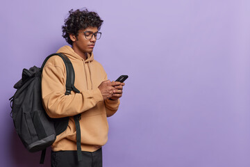 Horizontal shot of Hindu male college student messages on mobile phone while going home after classes dressed in casual hoodie carries black backpack isolated over purple background blank space