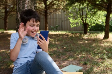 Cheerful teen boy waving hello with hand, having online conference by video link on his smartphone, sitting under a tree