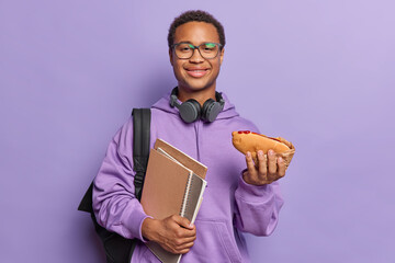 Pleased hungry male student holds hot dog and notepads wants to eat dressed in casual sweatshirt...