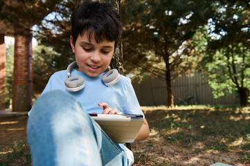 Smart teenage boy, primary school student with headphones around his neck, smiling, using digital tablet, doing homework