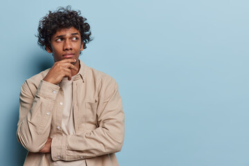 Horizontal shot of thoughtful curly haired Hindu man keeps hand on chin being deep in thoughts...