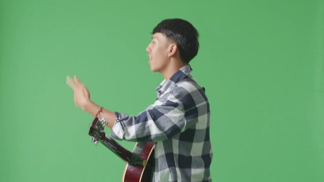 Side View Of Young Asian Teen Boy With Guitar And Rapping On The Green Screen Background

