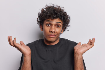 Young curly haired Hindu man in his 20s wears casual black t shirt expresses sense of confusion and...