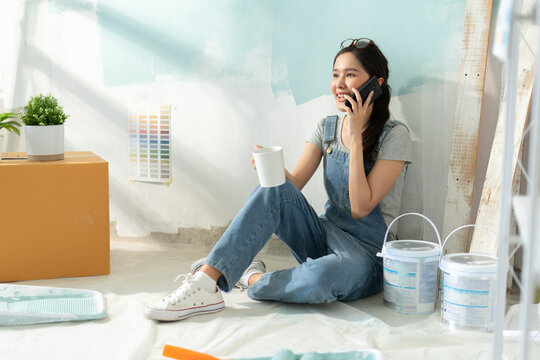 Smiling asian woman using phone after painting in room at new home.Asian young woman resting with coffee after work concept of paint color,moving house,new apartment,decorating house,new house.